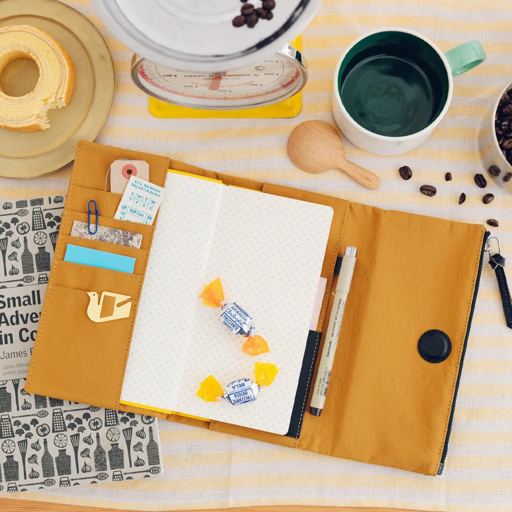 A mustard yellow fabric notebook cover laid open on a striped fabric surface, holding a white notebook, pens, clips, sticky notes, and small candies. Surrounding props include a coffee cup, scale, spoon, coffee beans, and a magazine.