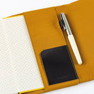 A mustard yellow fabric notebook cover shown open on a white background, with a white-and-silver pen tucked into a side pocket and a black leather card holder. 