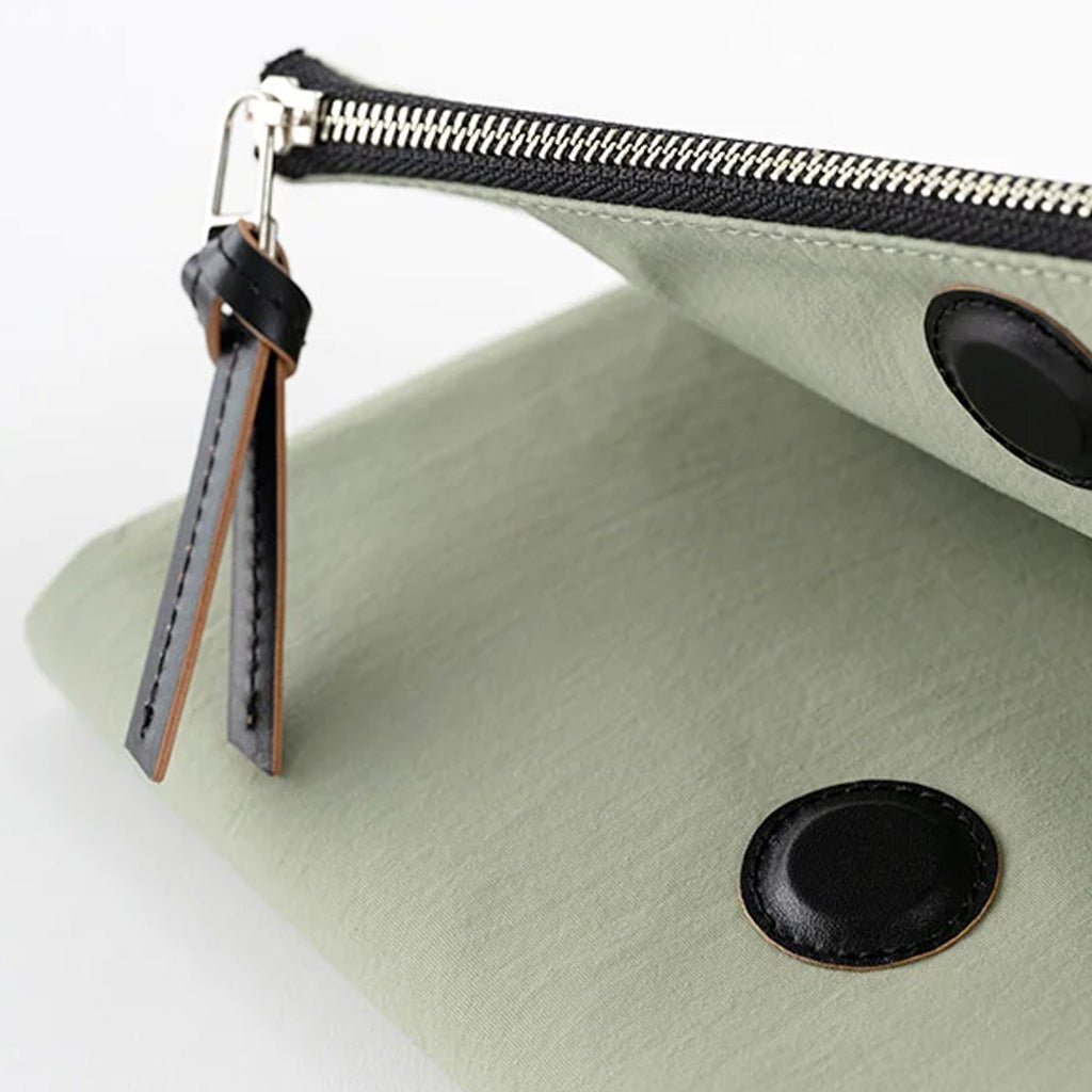 A close-up of a sage green cover with black leather trim, a black zipper, and a black leather pull tab with silver metal hardware, set against a clean white background.