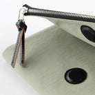A close-up of a sage green cover with black leather trim, a black zipper, and a black leather pull tab with silver metal hardware, set against a clean white background.