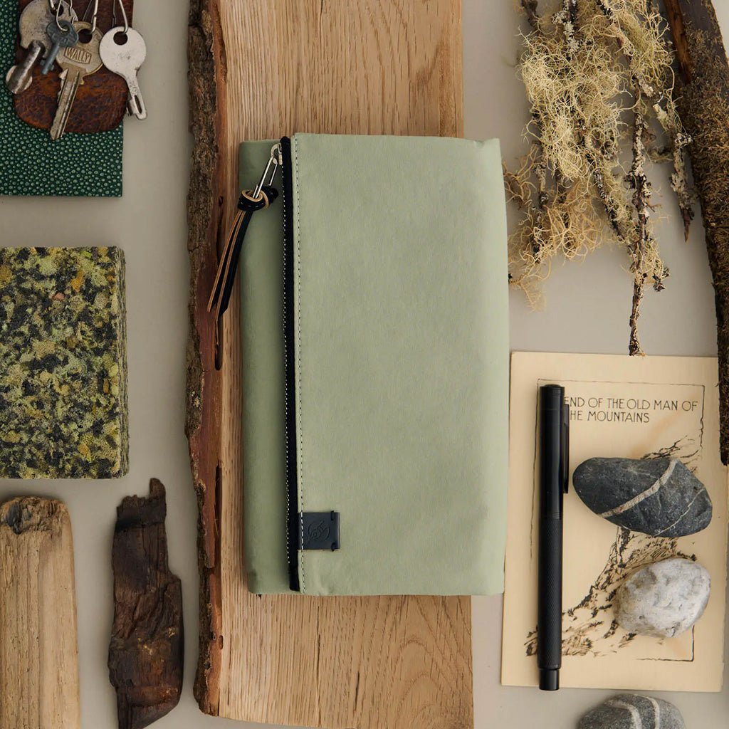 A sage green zippered pouch with a black zipper and leather pull tab, styled in a flat lay on a wood block surrounded by natural elements, keys, a notebook, and a pen.