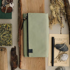 A sage green zippered pouch with a black zipper and leather pull tab, styled in a flat lay on a wood block surrounded by natural elements, keys, a notebook, and a pen.