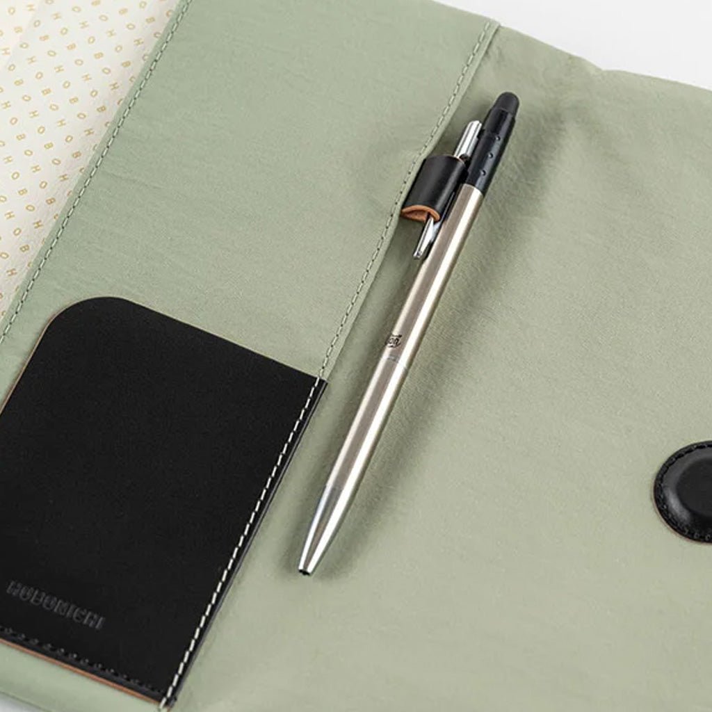 A silver and black pen resting on a light sage green fabric notebook cover with a black leather pocket on the left side.