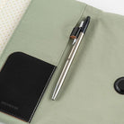 A silver and black pen resting on a light sage green fabric notebook cover with a black leather pocket on the left side.
