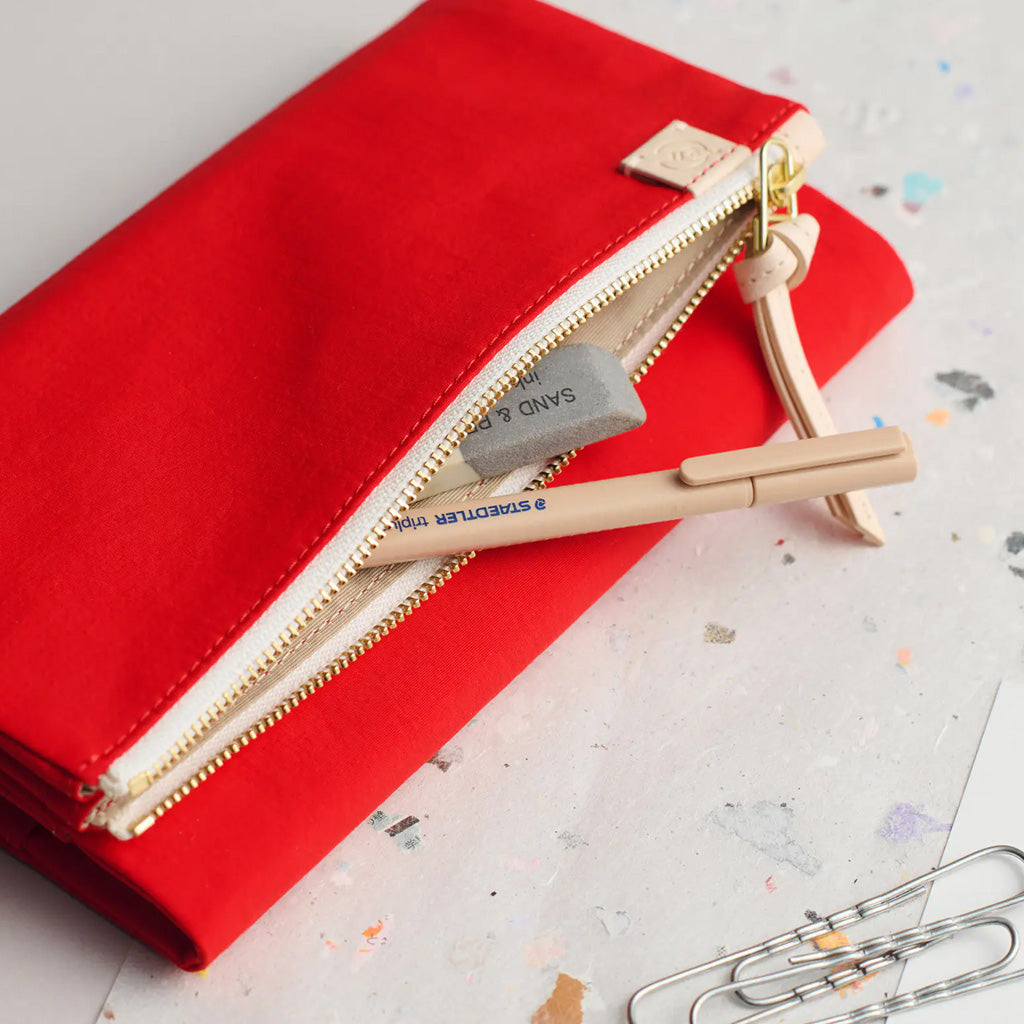 A red fabric pencil case with a cream zipper and pull tab, shown open on a speckled white surface. A gray eraser and a beige pencil are placed inside, with paperclips visible in the lower right corner.