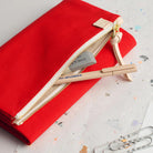 A red fabric pencil case with a cream zipper and pull tab, shown open on a speckled white surface. A gray eraser and a beige pencil are placed inside, with paperclips visible in the lower right corner.