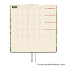 An open monthly calendar planner with cream grid pages, a floral-patterned cover visible along the edges, and a black ribbon bookmark. 