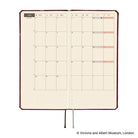 An open planner showing a monthly calendar spread on cream pages with a dark burgundy cover edge and ribbon bookmarks hanging below, photographed on a white background. 