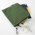 An olive green canvas laptop sleeve with a black zipper, shown open and styled with a notebook, pen, postcard, and white wired earphones on a clean white background.