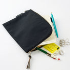A black zippered fabric pouch with a silver zipper and tassel, shown open with stationery items like a notebook, pen, ruler, and binder clips on a white background.