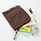 A brown zippered sleeve or pouch made of textured fabric, shown with stationery and tech accessories on a clean white background.
