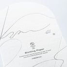 A close-up of a sheet of TSUKI no IRO Blotting Paper from the Hobonichi Techo series, featuring fine black line art, subtle gray abstract shapes, and two small blue-gray blotches on a white textured paper background.