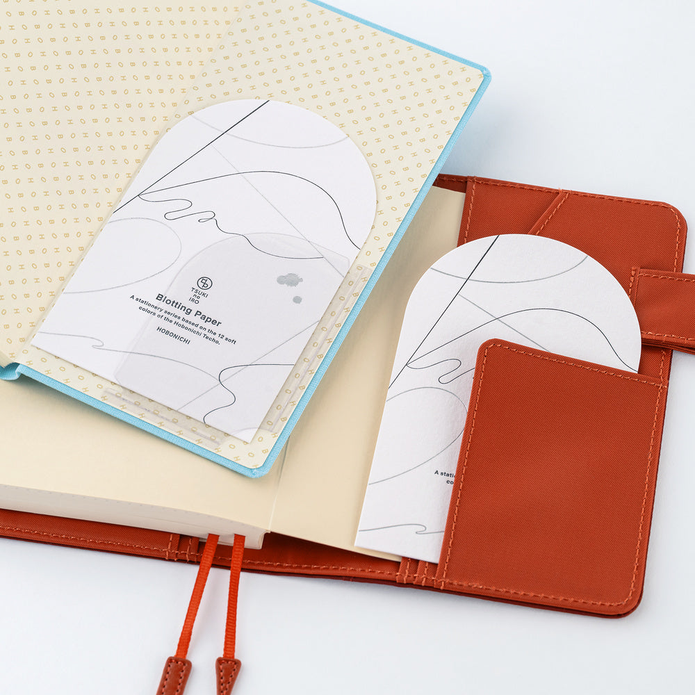 An open notebook with a light blue border and cream pages, featuring two white arched paper sheets with minimalist line art and the text 'Blotting Paper' in an orange-brown cover with a pocket.