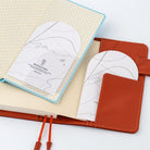 An open notebook with a light blue border and cream pages, featuring two white arched paper sheets with minimalist line art and the text 'Blotting Paper' in an orange-brown cover with a pocket.