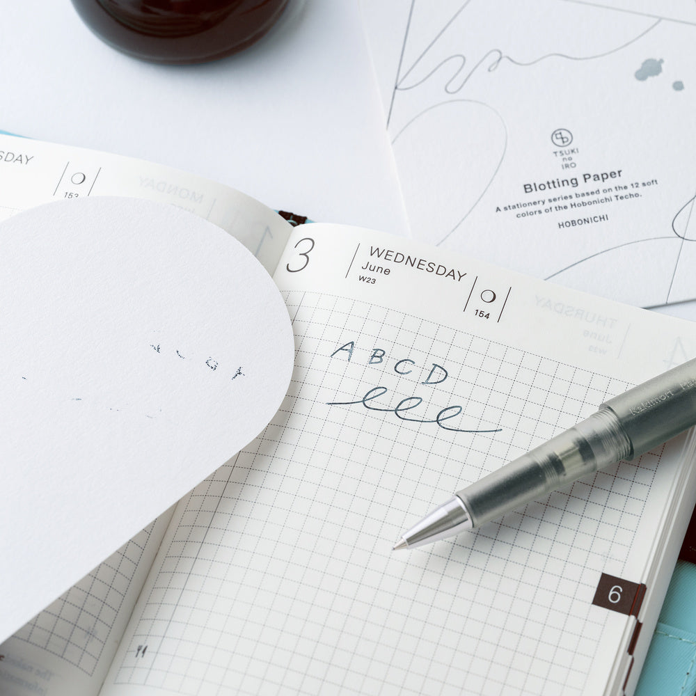An open grid planner or notebook on a white surface, with a translucent gray pen resting on the page and a sheet of blotting paper partially covering the left side. The visible text on the paper reads 'Blotting Paper' and 'Hobonichi'.