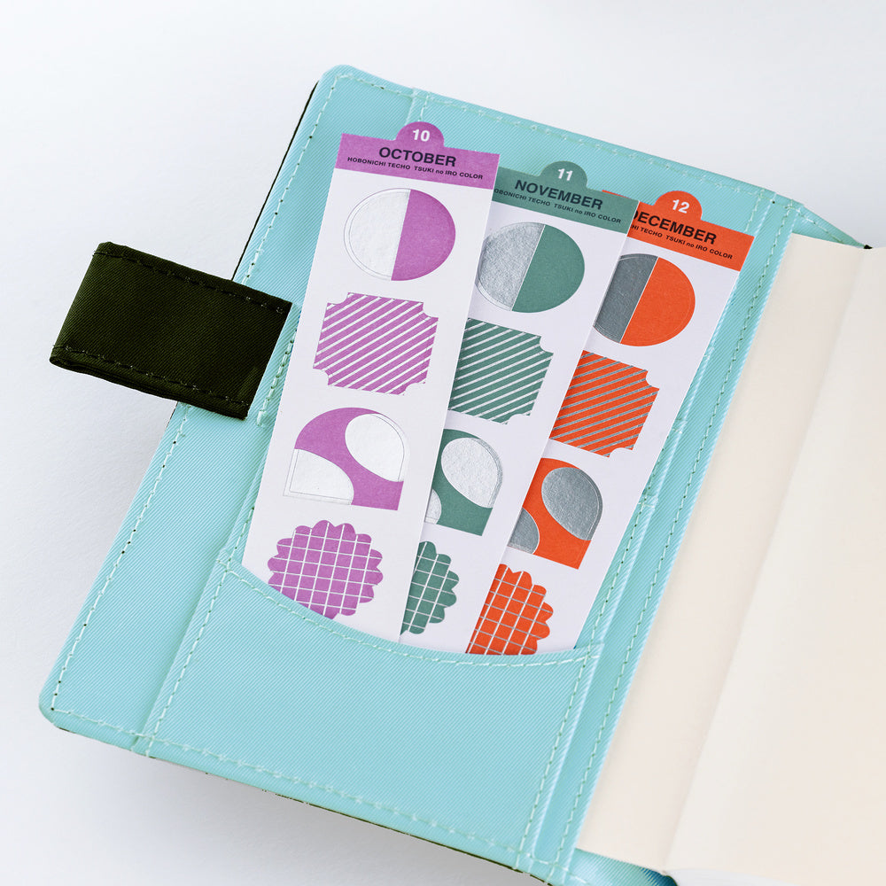 A light blue planner cover with a pocket holding two sticker sheets. The stickers feature geometric shapes in purple, green, orange, and gray, labeled for October, November, and December.