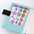 A light blue planner cover with a pocket holding two sticker sheets. The stickers feature geometric shapes in purple, green, orange, and gray, labeled for October, November, and December.