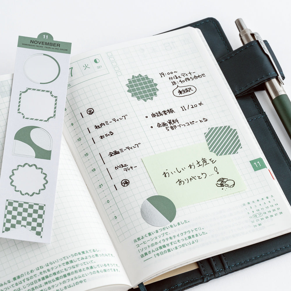 An open planner or notebook with Japanese writing and green patterned accents, shown alongside a matching planner sticker sheet and a pen on a white background.