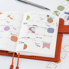 An open grid planner or bullet journal on an orange faux-leather cover, styled on a white surface with a pen and patterned paper sheets nearby. The planner pages show decorated monthly calendar layouts with pastel and earth-tone stickers.