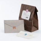 A brown paper gift bag with a white note card, a light gray envelope with a small orange patterned tab, and a cream-colored card with a blue patterned tab, arranged on a clean white background.