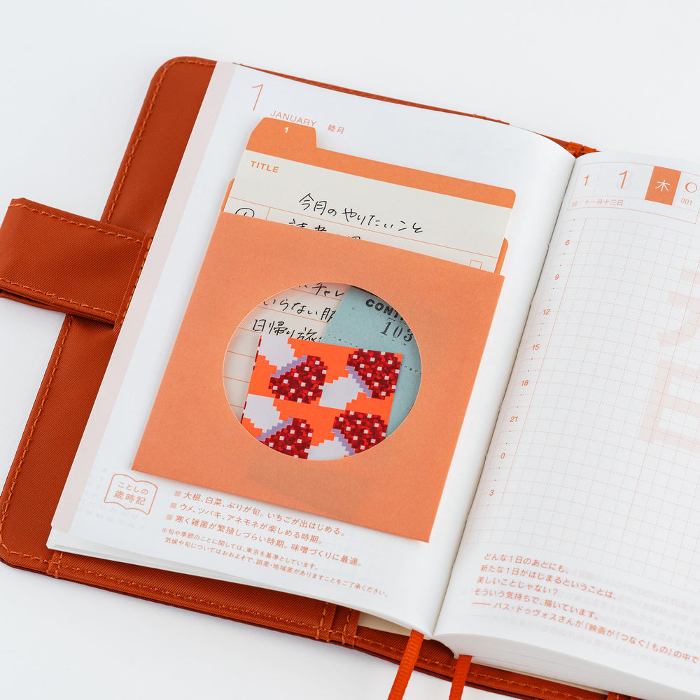An open planner with Japanese text and grid pages, featuring a paper pocket with a circular cutout revealing a colorful patterned insert.