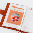 An open planner with Japanese text and grid pages, featuring a paper pocket with a circular cutout revealing a colorful patterned insert.