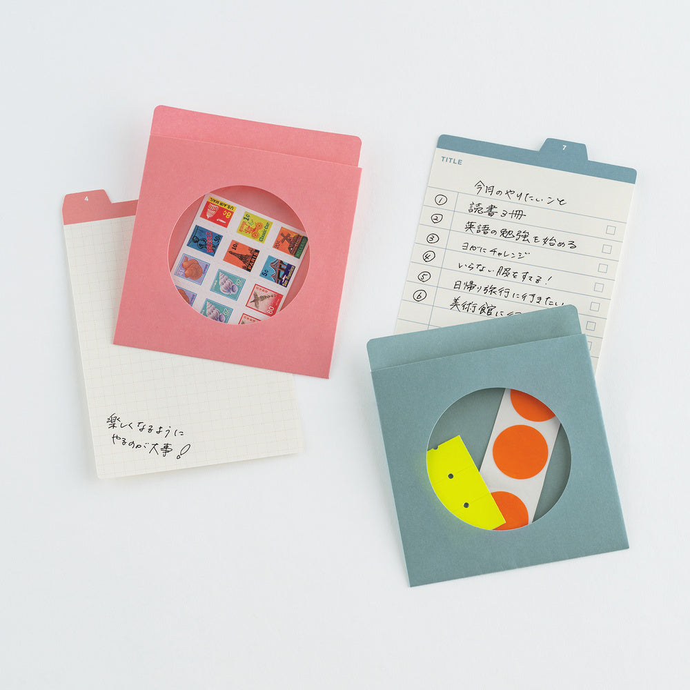 Three colorful paper envelope-style stationery pieces arranged on a white background: a pink envelope with a circular cutout showing a sheet of small illustrated stickers, a white grid notepad with handwritten text, and a muted blue envelope with a circular cutout showing a sheet of orange and yellow dots.