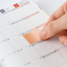 A close-up of a hand applying a peach-colored adhesive sticky note to a white grid calendar planner.
