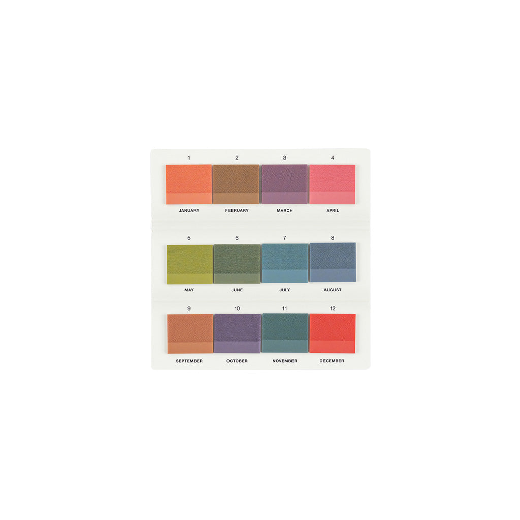 A white rectangular card displaying twelve monthly colored sticky notes arranged in a 3x4 grid, labeled January through December with corresponding pastel and muted color blocks.