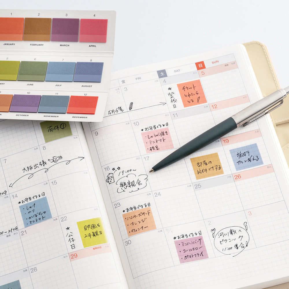 An open planner showing a monthly calendar spread with handwritten notes, colorful sticky notes, a pen, and a small color card sheet on a clean white background.
