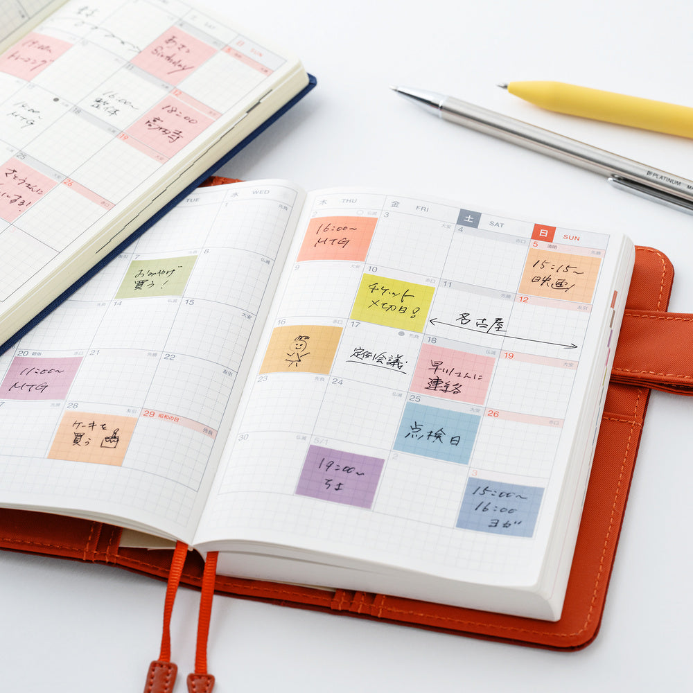 An open orange-brown planner with a daily calendar layout and colorful sticky notes, photographed on a white surface with other stationery items nearby.