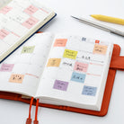 An open orange-brown planner with a daily calendar layout and colorful sticky notes, photographed on a white surface with other stationery items nearby.