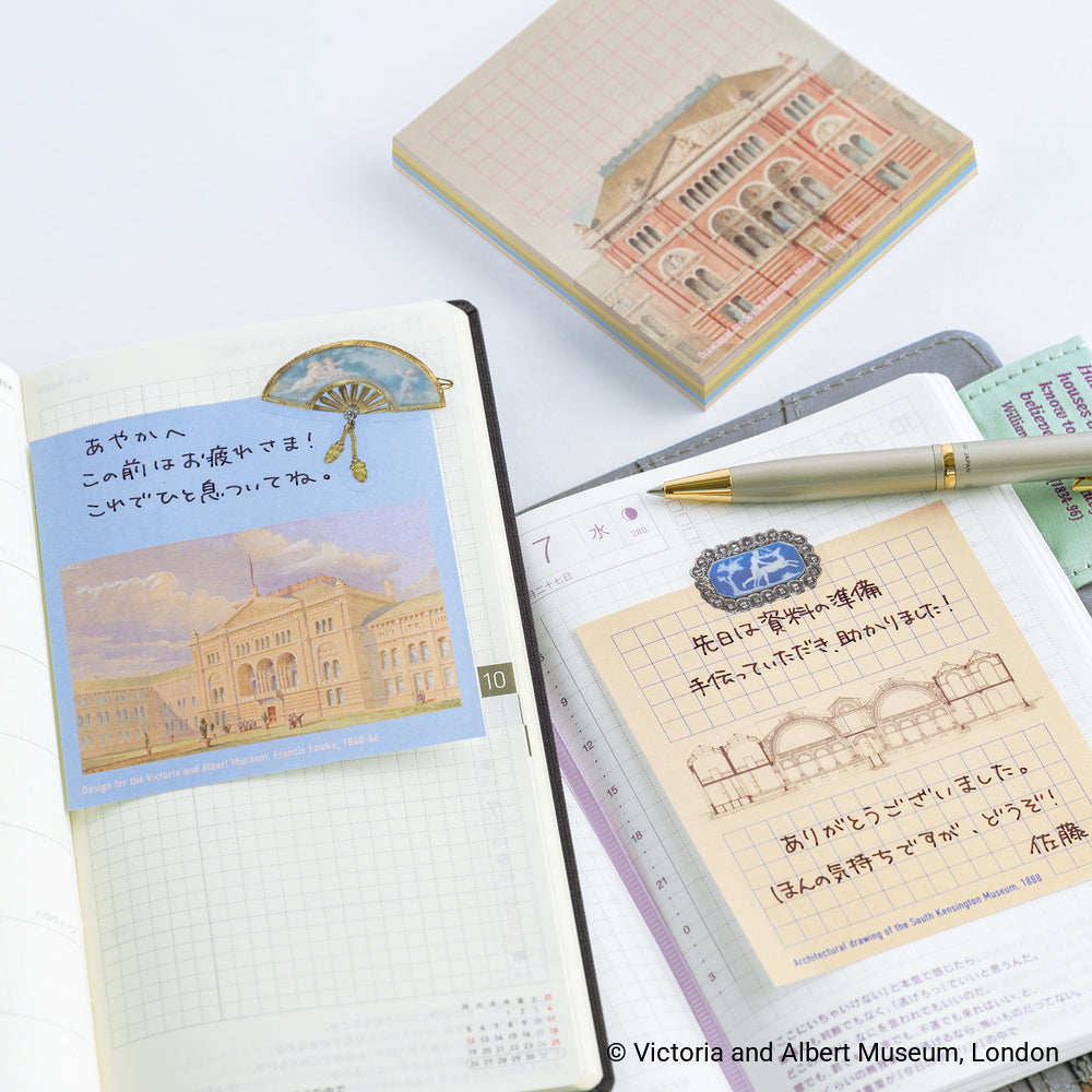 A styled flat-lay of stationery featuring a Victoria and Albert Museum themed notepad, sticky notes, and a gold pen on open grid notebooks with Japanese writing.