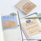 A styled flat-lay of stationery featuring a Victoria and Albert Museum themed notepad, sticky notes, and a gold pen on open grid notebooks with Japanese writing.