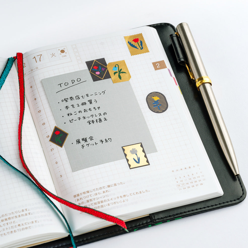 An open black notebook with a grid page, decorated with Yoshie Watanabe stickers with gold foiling and colorful flower motifs alongside a metallic pen on a white background.