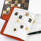 An open orange-brown planner with a grid calendar page, decorated with Yoshie Watanabe stickers with gold foiling and colorful flower motifs. A black pen rests on the page, and a sheet of matching decorative stickers is visible in the background on a white surface.