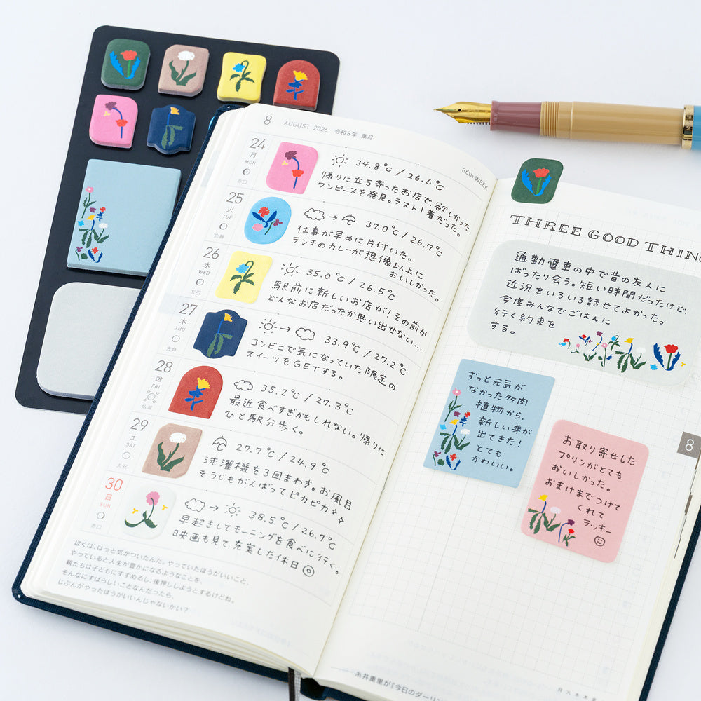 An open planner with Japanese notes and colorful floral sticky notes, shown alongside a black notepad set with matching floral tabs and a gold pen on a white background.