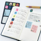 An open planner with Japanese notes and colorful floral sticky notes, shown alongside a black notepad set with matching floral tabs and a gold pen on a white background.