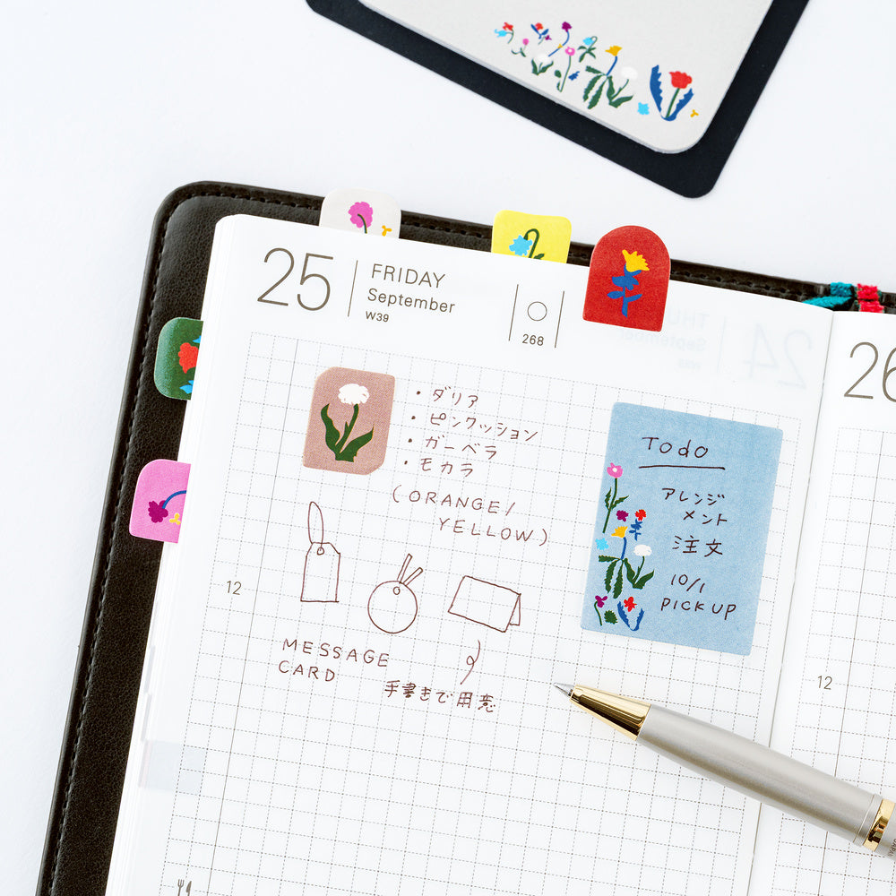 A styled flat-lay of a planner open to a daily page, decorated with colorful floral sticky notes, and a pen. The scene is photographed on a clean white background.
