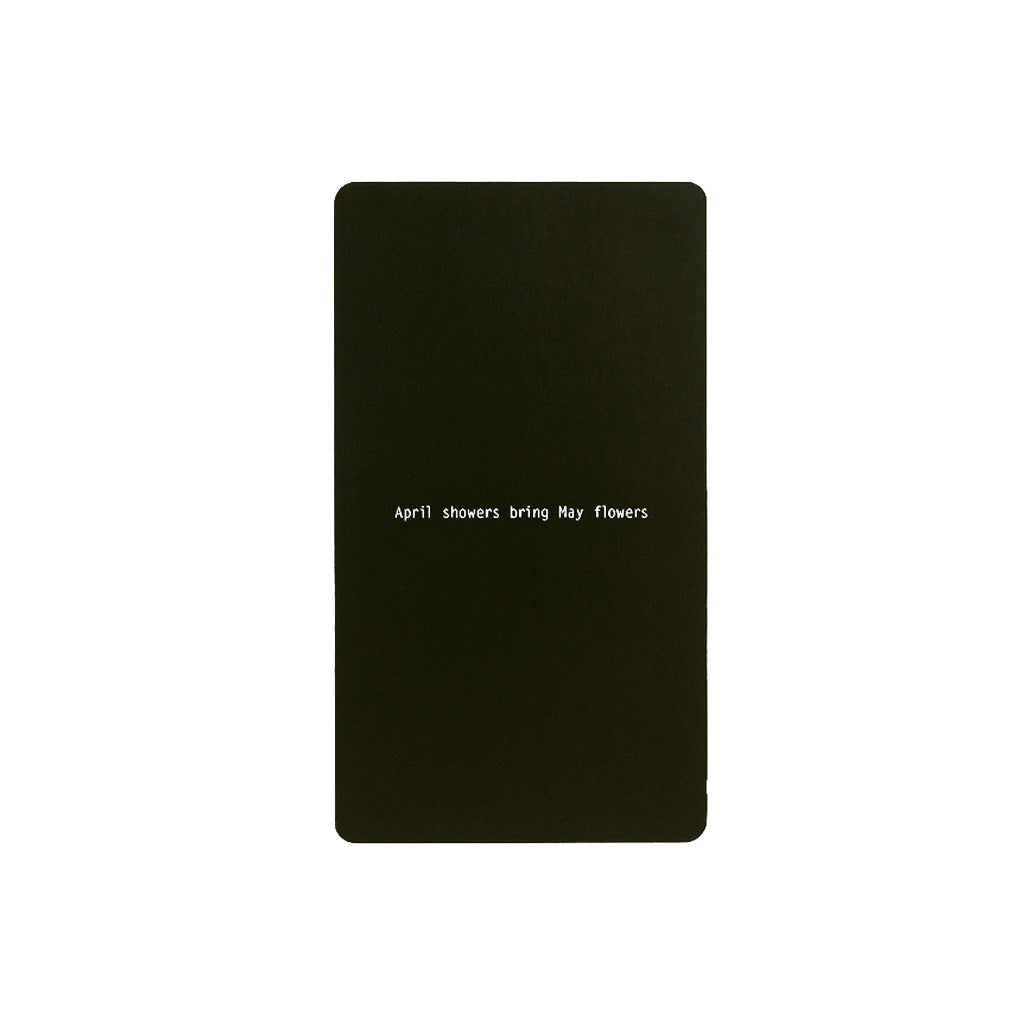 A black rectangular notepad with rounded corners, featuring the phrase 'April showers Bring May flowers' printed in small white text in the center, set against a plain white background.