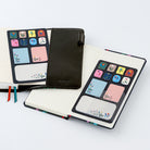 A black folio-style planner cover with a clear pocket displaying colorful decorative sticky notes shown alongside an open notebook on a clean white background.