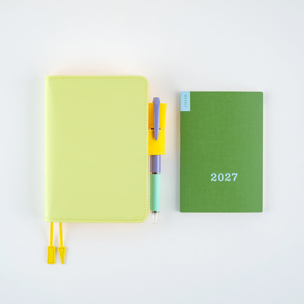 A flat-lay arrangement on a white background featuring a pale yellow planner with a yellow bookmark ribbon, a pastel pen in a yellow and purple sleeve, and a green 2027 weekly planner.