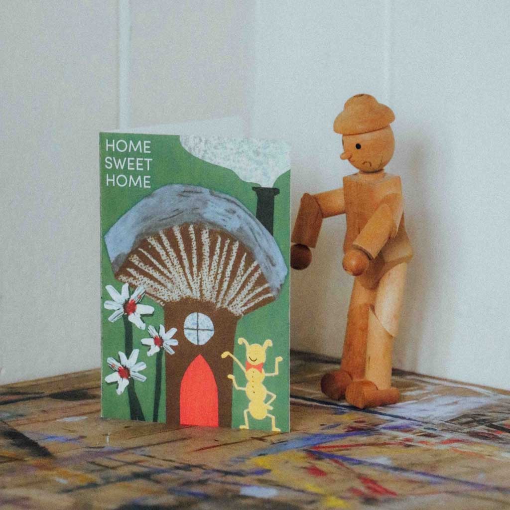 Wooden figurine standing next to a 'Home Sweet Home' card with a house illustration on a textured surface.