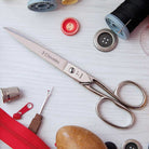 Sewing scissors with '3 Claveles' branding on a white surface with sewing accessories.