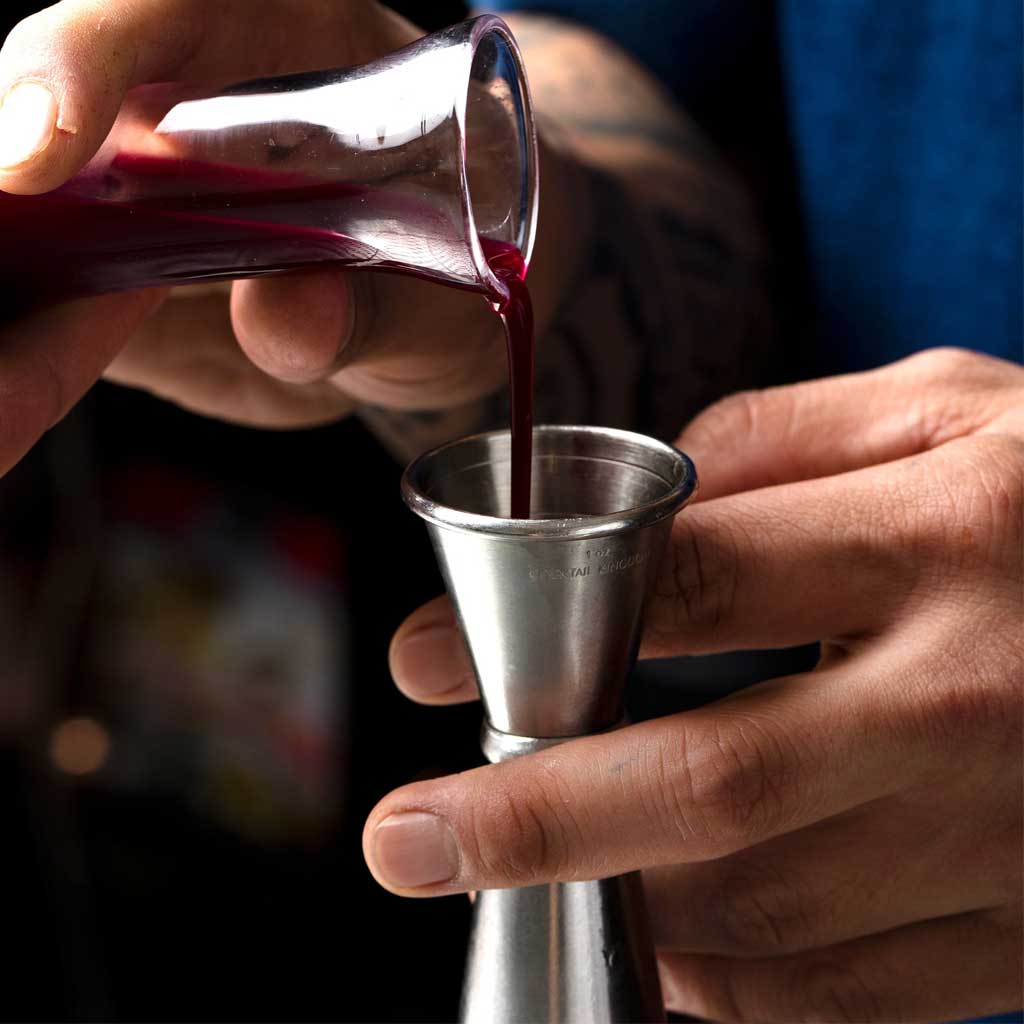 Person pouring red liquid from a glass into a metal jigger.