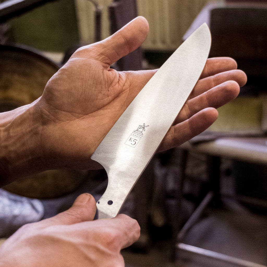 Hand holding a large k5 knife blade with a blurred workshop background