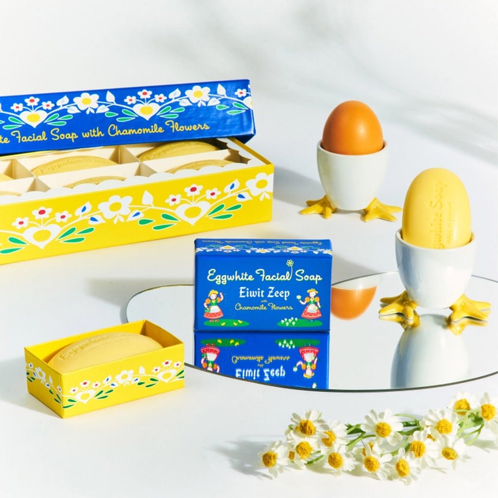 Eggwhite Facial Soap in open box in front of soap gift box set around mirror and egg cups against light background