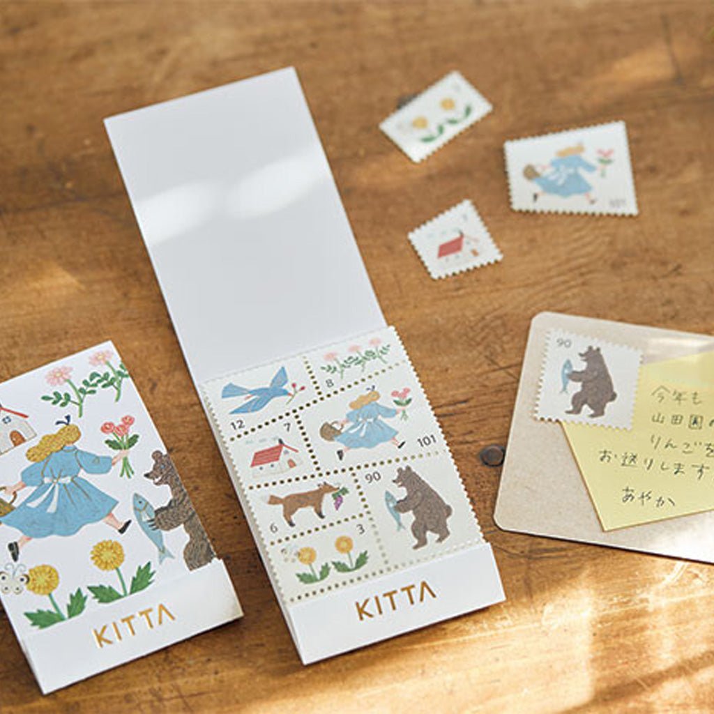 Set of washi stamp-style stickers in paper holder with gold "KITTA" branding on bottom, surrounded by stationery on wooden surface