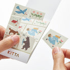Hand holding stamp-style washi sticker with colorful illustration of girl running next to set of stickers in holder against white background