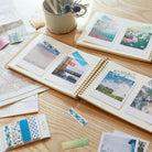 Open photo album with scenic photos taped with washi tape on a wooden surface with decorative items.